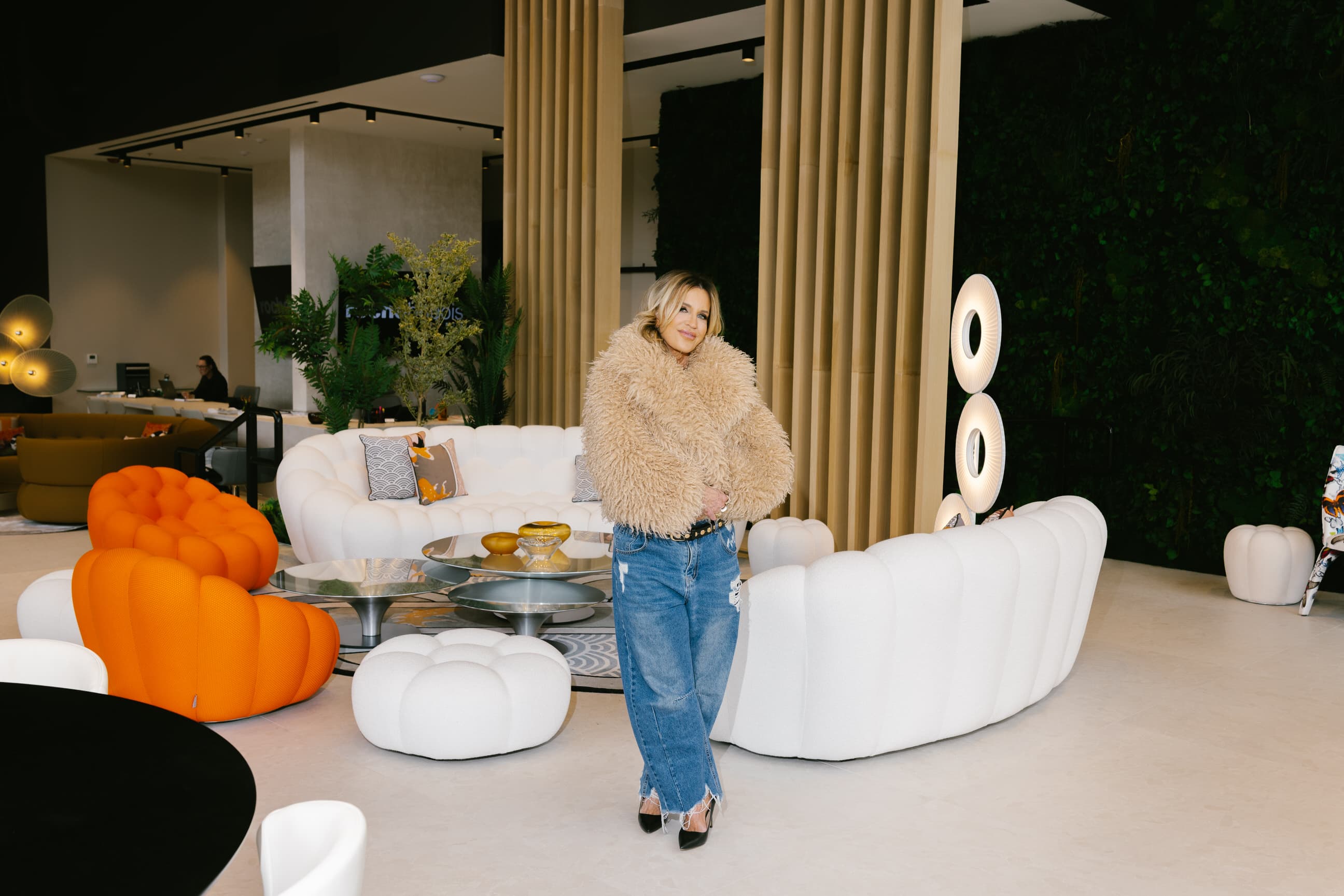 Giselle Koy in a tan shearling coat among ivory bubble sofas and bamboo columns in a designed showroom