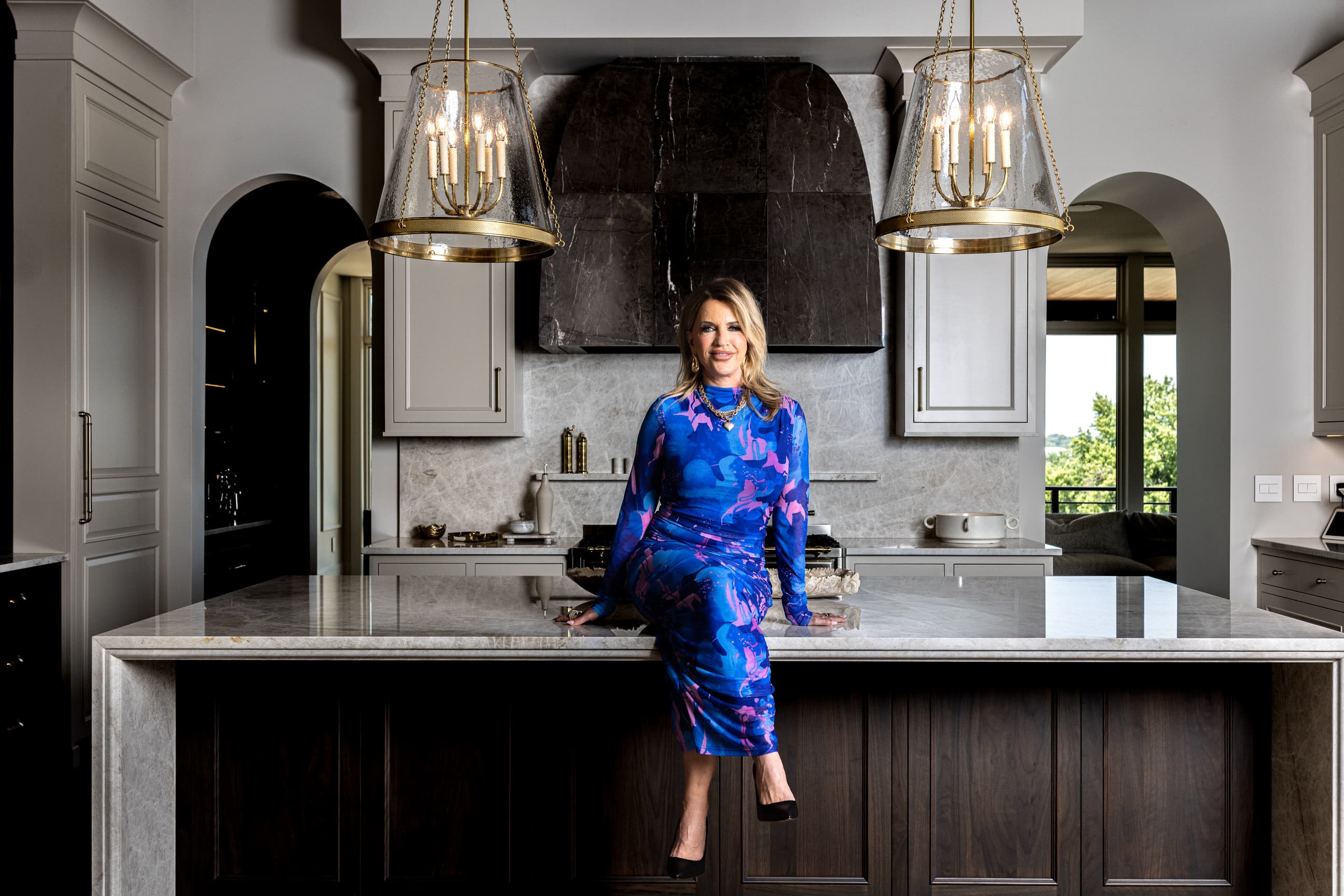 Giselle Koy seated on a Helmüt kitchen island, with gray upper cabinetry, marble countertops, and brass lantern pendants.