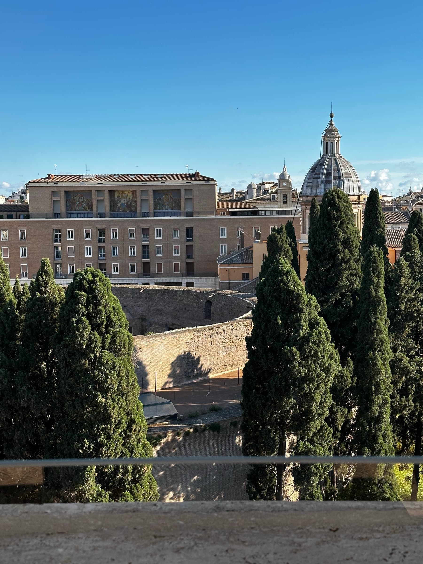 View from a terrace over the circular brick drum of the Mausoleum of Augustus, framed by cypress trees, with a church dome beyond.