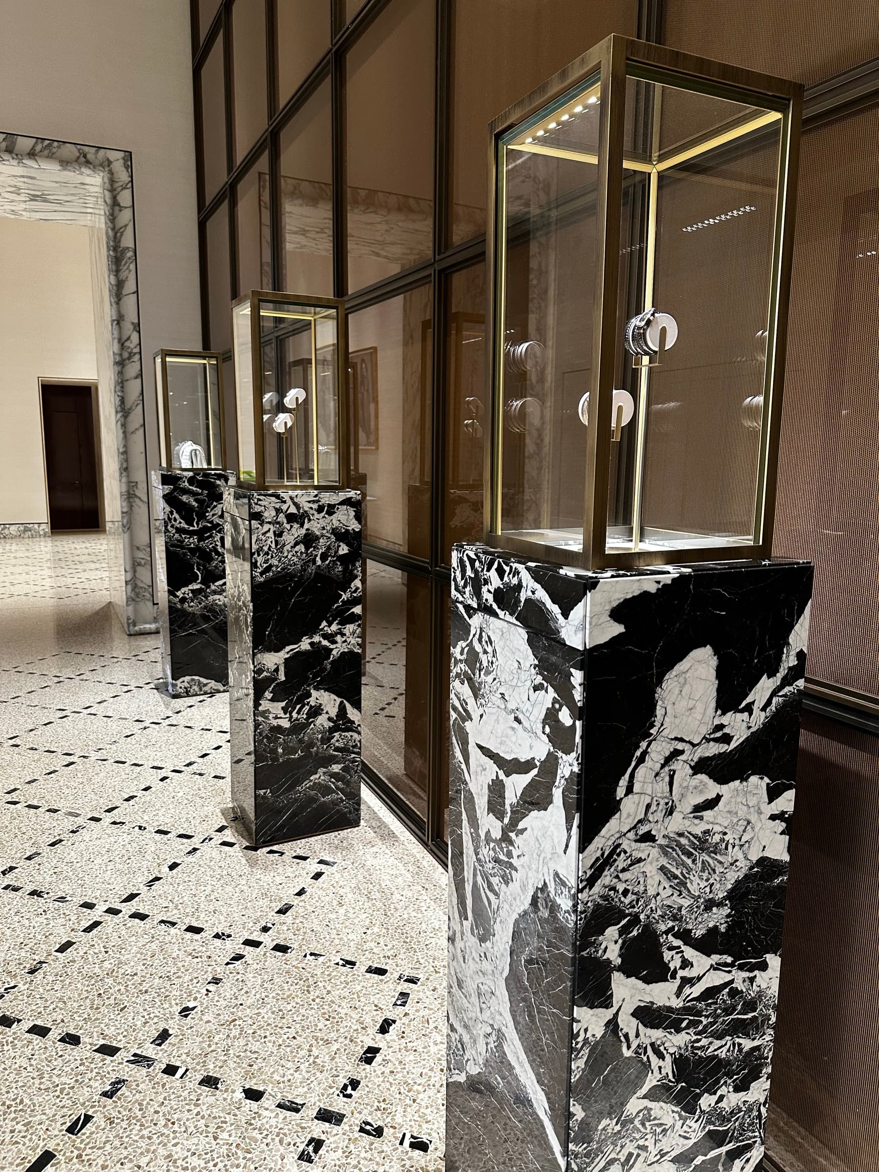 Three plinths of black marble veined in white, each topped with a brass and glass vitrine holding jewelry, on an inlaid terrazzo floor.