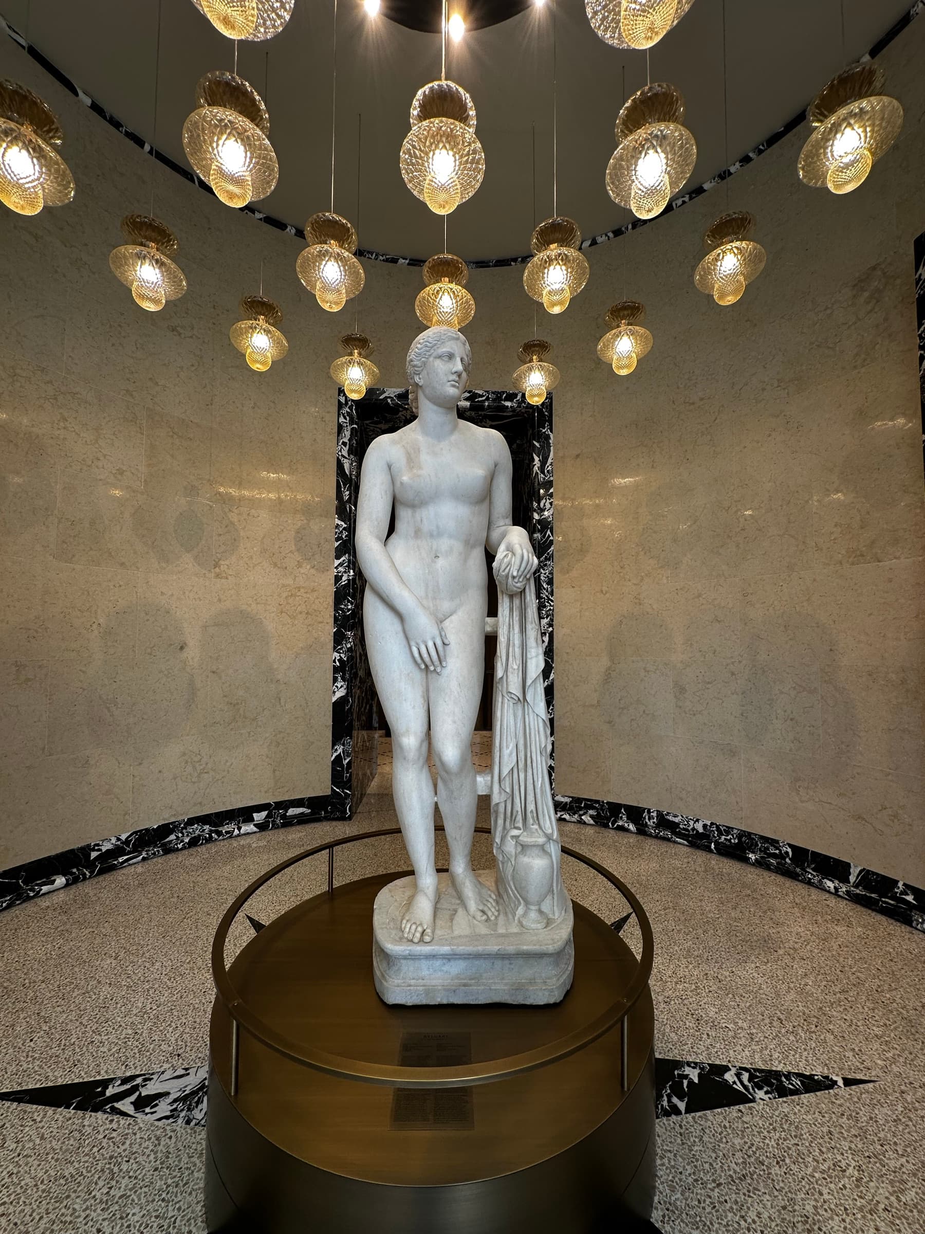 A classical marble figure on a round plinth beneath a cluster of golden glass pendant lights, against a panel of black and white marble.