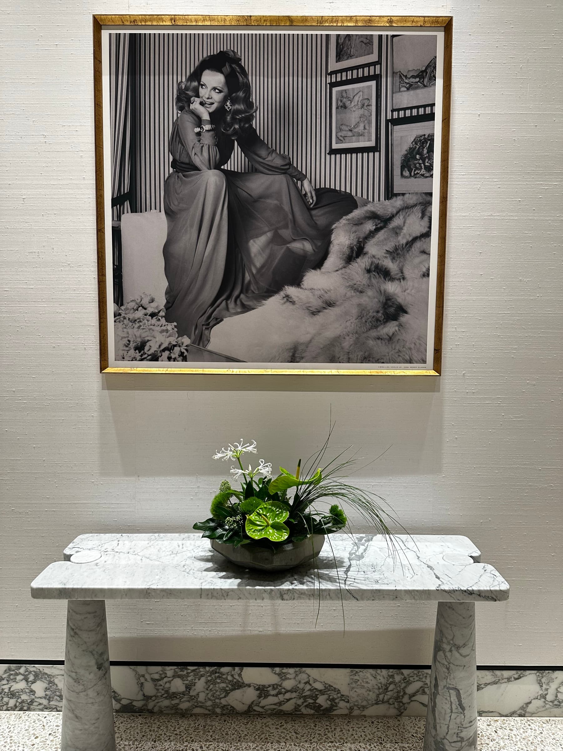 A framed black and white photograph in a gold frame above a white marble console table with a green floral arrangement.