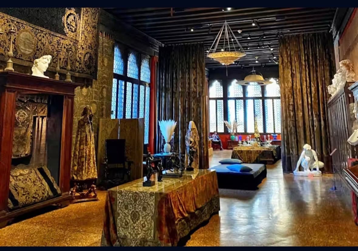 The grand salon of Palazzo Fortuny — gold and bronze Fortuny fabric draped across tables, arched Gothic windows, and dark ceiling beams.