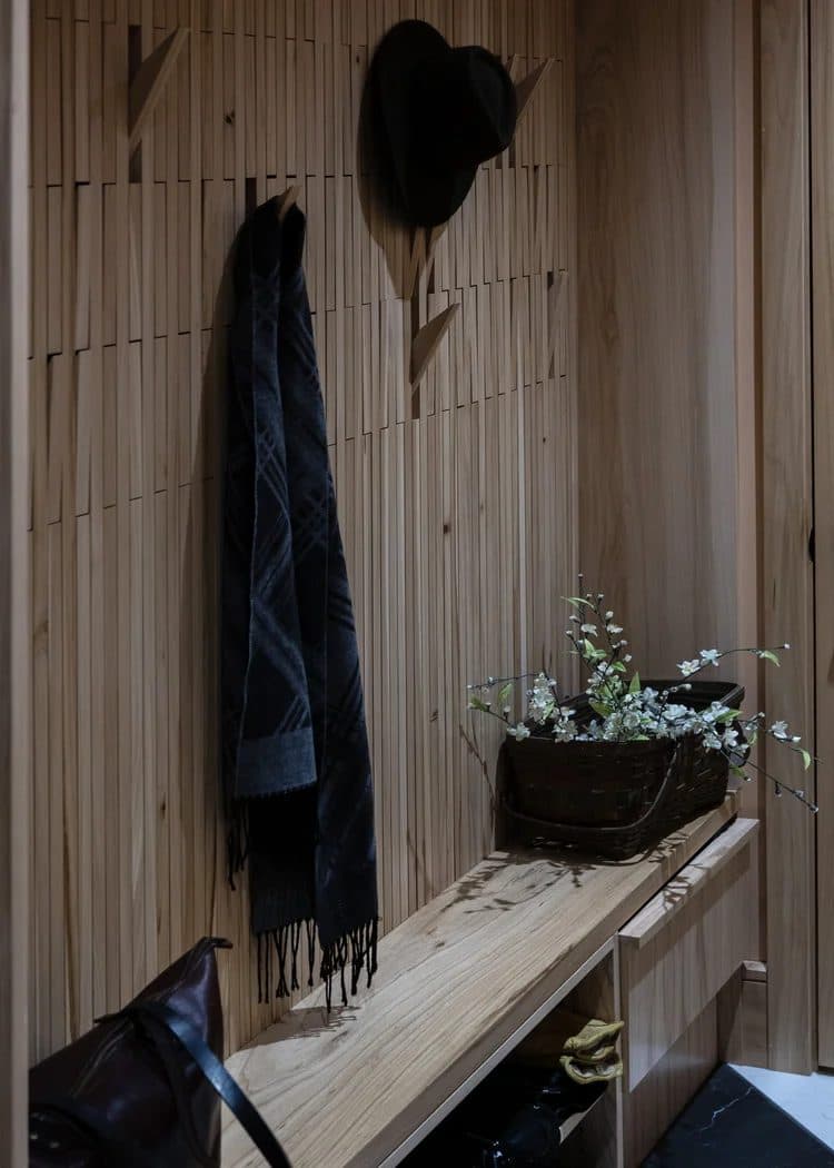 Natural wood slatted mudroom entry with built-in bench and coat hooks holding a jacket and hat.
