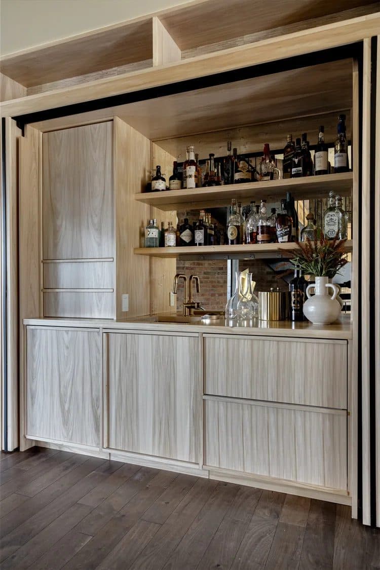 Light wood bar cabinet with sliding doors open to reveal shelved bottles, glassware, and a vase of flowers.