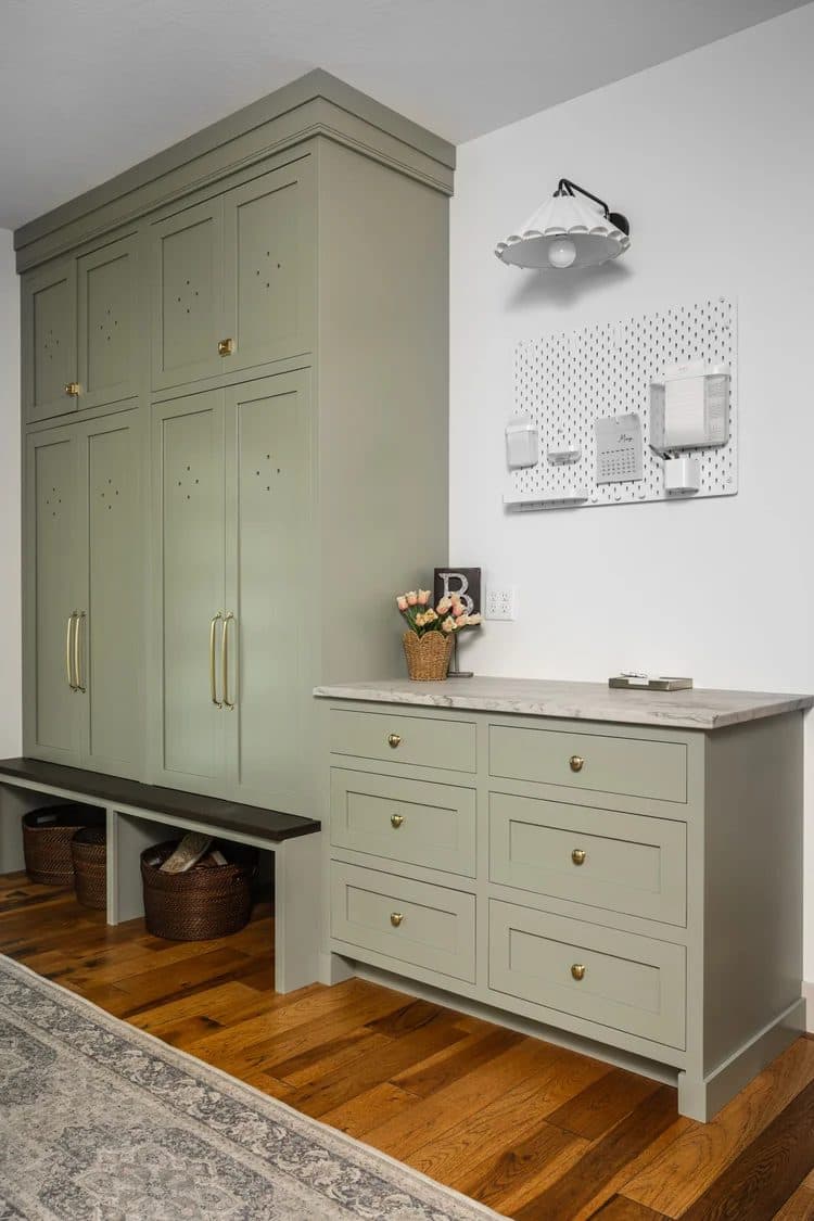 Sage green painted mudroom cabinetry floor-to-ceiling with brass hardware, bench, and drawer unit on warm wood floors.