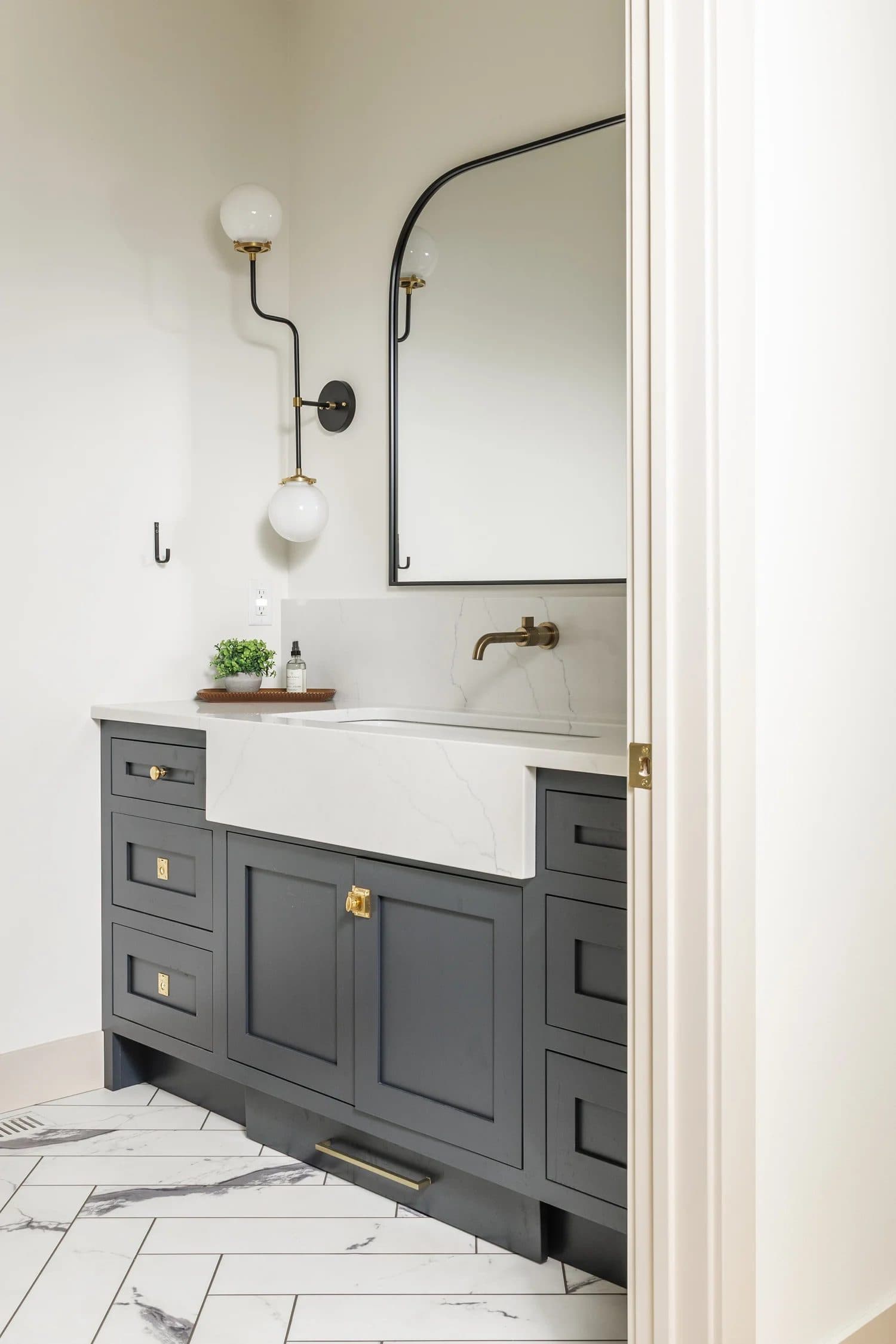 Charcoal bathroom vanity with farmhouse sink, arched mirror, wall sconce, brass hardware, and herringbone marble floor.