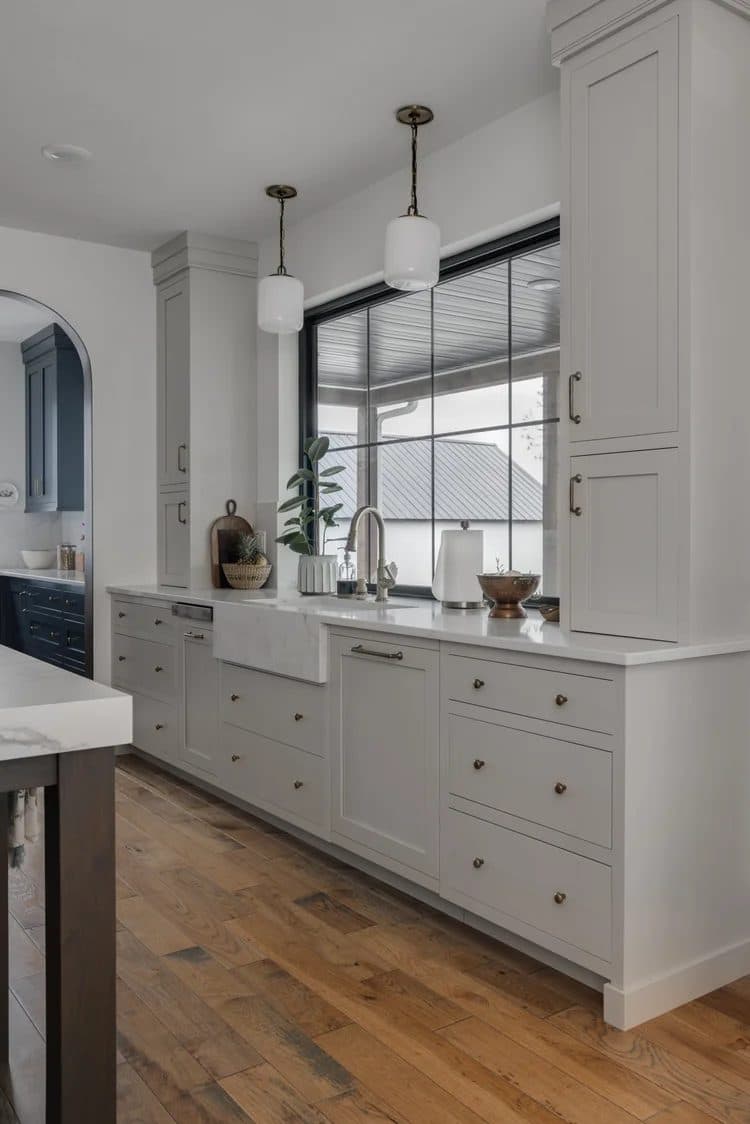 Cream kitchen with farmhouse sink, white shaker cabinetry, wood floors, and pendant lights over an island.