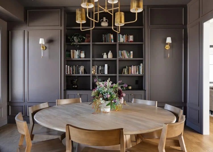 Dark gray dining room with built-in shelving, round wood dining table, chairs, and gold chandelier.