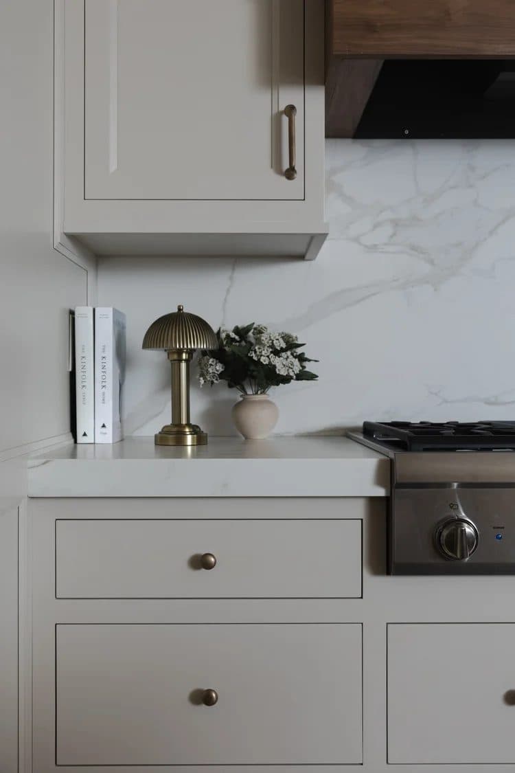 Detail of cream kitchen cabinetry with brass hardware, marble backsplash, and a brass lamp beside the range.