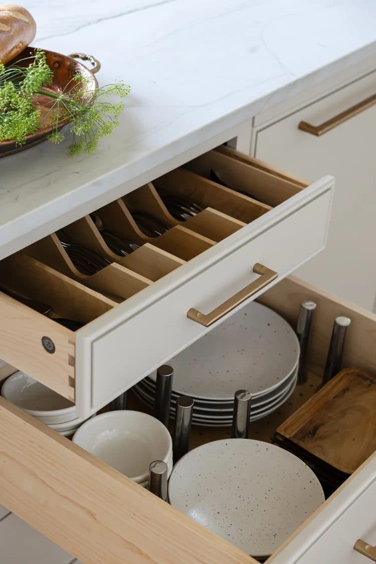 Open kitchen drawer with custom wood utensil dividers, stacked plates and bowls visible in the drawer below.