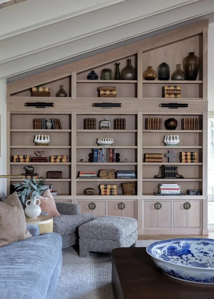 Natural wood built-in bookshelves filling a sloped ceiling alcove, loaded with books, ceramics, and objects.